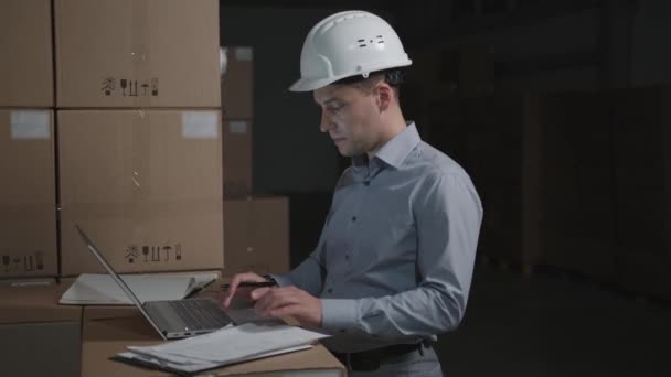 El hombre en el almacén trabaja con el portátil sobre el fondo de las cajas de cartón — Vídeos de Stock