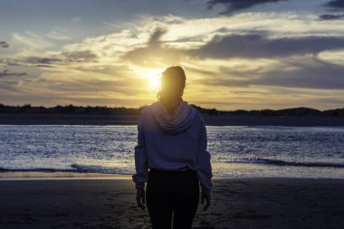 Sahilde genç bir atlet kadın gün batımını izliyor, onun siluetini görebilirsiniz ve arkadan poz veriyor.