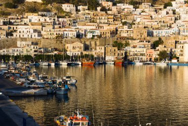 Kalymnos Adası Yunanistan. Şafak vakti, körfezi ve limanı çevreleyen zarif neo klasik evlerin üzerinde ışıldayan yumuşak güneş ışığıyla birlikte. Manzaralı körfez ve limanda küçük tekneler var..