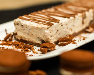 Uzun beyaz bir tabakta çikolata soslu dikdörtgen peynirli kek ve etrafında beyaz kremalı çikolatalı kurabiye.