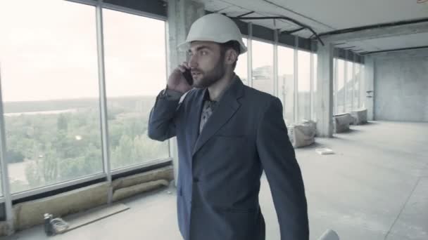 El joven arquitecto pasa por las instalaciones del edificio reparado, hablando en el teléfono inteligente — Vídeo de stock