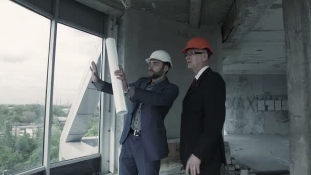 Dos hombres con traje y casco discuten el plano, calculan y se comunican con su colega. Americano — Vídeos de Stock