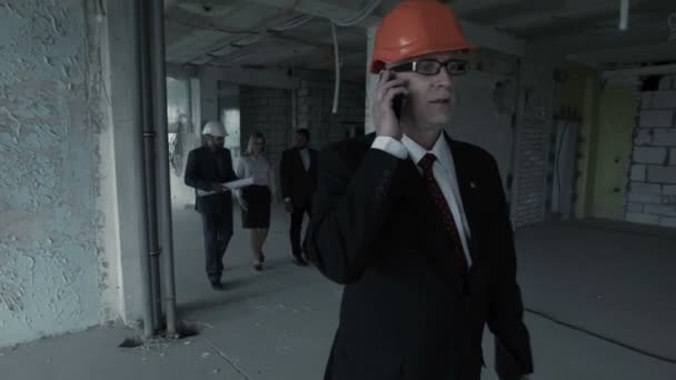 Man in suit, helmet call inside construction site. At background team of young builders man, african — Stock Video