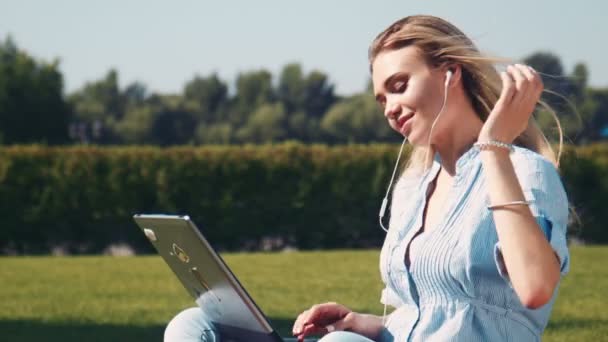 Çimenlerin üzerinde kulaklık dizüstü bilgisayar kullanan gülümseyen kız — Stok video