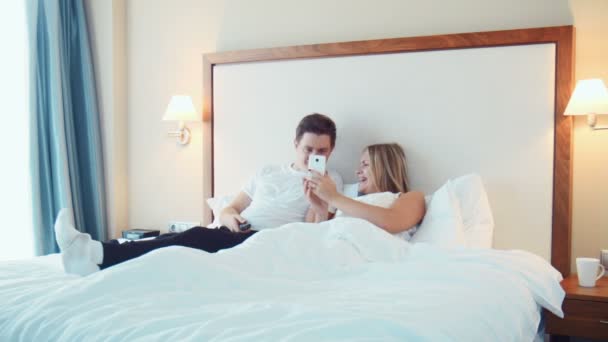 Pareja feliz tumbada en la cama viendo la televisión y el uso de smartphone — Vídeo de stock