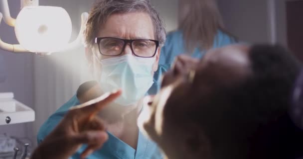 Зрілий стоматолог лікар вивчає зуби етнічного пацієнта — стокове відео