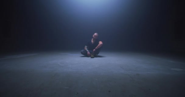 Tetovaný plešatý chlap sedí na podlaze v tmavé místnosti — Stock video