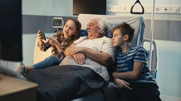 Paziente anziano che discute di video su smartphone con nipoti nel reparto ospedaliero — Foto Stock
