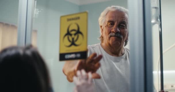 Starszy mężczyzna dotykający dłoni wnuczki za szklaną ścianą na oddziale COVID — Wideo stockowe