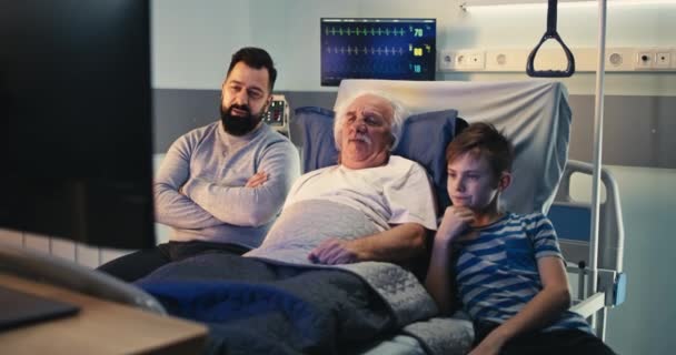 Пожилой мужчина празднует победу любимой команды с сыном и внуком в палате больницы — стоковое видео