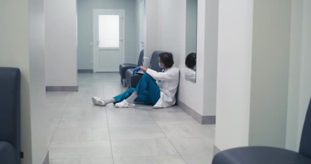 Gestresste dokter van middelbare leeftijd rustend in de hal van de kliniek tijdens een pandemie — Stockvideo