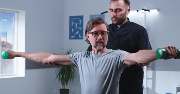 Terapeuta masculino que ayuda al paciente maduro a hacer ejercicio con pesas — Vídeo de stock