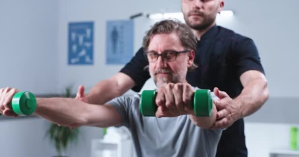 Střídavý terapeut pomáhá dospělému muži cvičit s činkami — Stock video