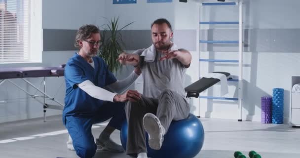 Terapeuta w średnim wieku pomaga pacjentowi utrzymać równowagę na piłeczce — Wideo stockowe