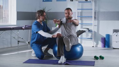 Orta yaşlı terapist hastanın topun dengelenmesine yardım ediyor.