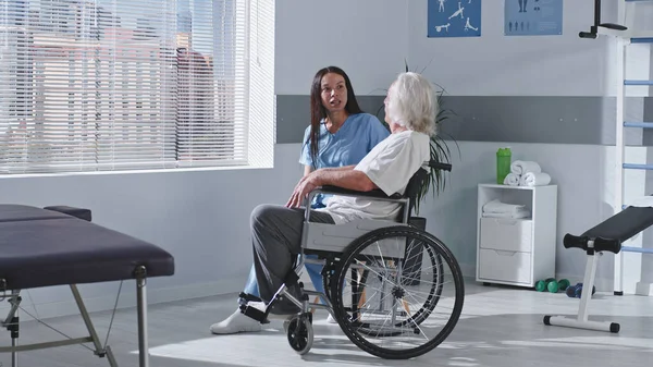 Amistoso terapeuta de rehabilitación hablando con un paciente anciano discapacitado — Foto de Stock