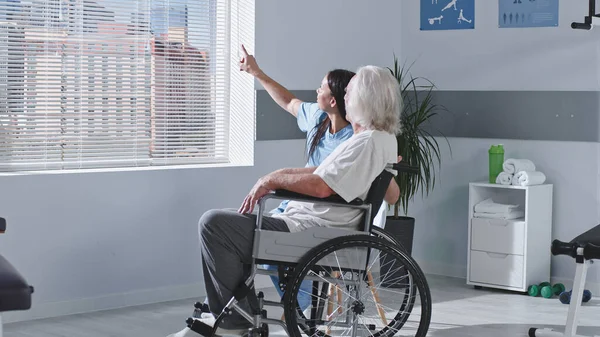 Amistoso terapeuta de rehabilitación hablando con un paciente anciano discapacitado — Foto de Stock