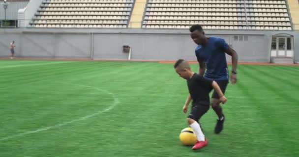 Черный тренер учит мальчика играть с мячом на футбольном поле — стоковое видео