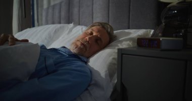 Olgun bir adam sabahın erken saatlerinde parlak güneş ışığıyla dolu bir odada uyanır. Dijital Alarm Saati 7: 00 'yi gösteriyor. Yavaş Sabah, Yatma Zamanı. Sabah rutini kavramı, günaydın. Sabit Çekim.