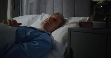 Orta yaşlı bir adam güneş ışığıyla dolu yatak odasında sabahın köründe uyanıyor. Gri saçlı adam yatakta uzanıyor, gözlük takıyor. Konsept Sabah Rutini, Rahatlama, Rahatlama, Konforlu Sabah.