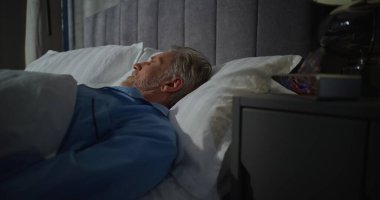 Olgun bir adam sabahın erken saatlerinde parlak güneş ışığıyla dolu bir odada uyanır. Dijital Alarm Saati 7: 00 'yi gösteriyor. Yavaş Sabah, Yatma Zamanı. Sabah rutini kavramı, günaydın. Sabit Çekim.