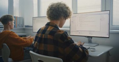 Two Young Men Writing Code in Classroom. University Students Sit at Computers, Attentively Typing Lines of Code During Programming Lesson. Concept of Teamwork, Higher Education. Back View. Static Shot