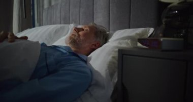 Olgun bir adam sabahın erken saatlerinde parlak güneş ışığıyla dolu bir odada uyanır. Dijital Alarm Saati 7: 00 'yi gösteriyor. Yavaş Sabah, Yatma Zamanı. Sabah rutini kavramı, günaydın. Sabit Çekim.