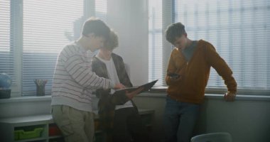 Pencere kenarındaki Parlak Sınıfta Üç Öğrenci Konuşuyor. Smartphone 'da Üçüncü Harcama Zamanı' nda Oyun Geliştirme Projesi İncelemesi Hakkında Etkin Konuşan İki Adam. Öğrenci yaşam tarzı. Yavaş Hareket.