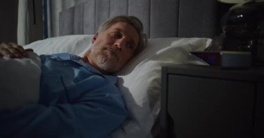 Orta yaşlı bir adam güneş ışığıyla dolu yatak odasında sabahın köründe uyanıyor. Gri saçlı adam yatakta uzanıyor, gözlük takıyor. Konsept Sabah Rutini, Rahatlama, Rahatlama, Konforlu Sabah.