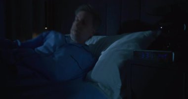 Mavi Pijamalı Adam Yatağa Gidiyor, Dijital Alarm Saati Sabahın 3 'ünü gösteriyor. Uykusuzluktan yorgun düşen adam sabahın erken saatlerinde uyumaya çalışır. Uykusuzluk kavramı, gece endişesi ile sessiz mücadele..