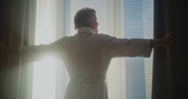 Olgun adam pencereden dışarı bakar, ağır perdeleri geri çeker ve gerilir. Parlak güneş ışığıyla dolu modern bir otel odası. Rahatlama, seyahat, günaydın, duygusal refah kavramı. Arka Görünüm.