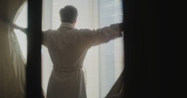 Son sınıf öğrencisi, Büyük Otel Pencerelerinden Sabah Güneşinin İlk Işıkları 'nın keyfini çıkarıyor. Adam Perdeleri ve Esnemeleri Geri Çekiyor. İyi Varoluş, Lüks Seyahat Yaşam Tarzı ve Düşünceli Sabah Enerjisi kavramı. El bilgisayarı.