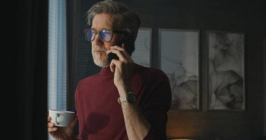 Modern Otel Odası 'ndaki zarif gri saçlı adam elinde kahve fincanı tutuyor ve Smartphone' da konuşuyor, Pencereden Manzaranın tadını çıkarıyor. Rahat İş Sabahı kavramı, Dijital Bağlantı, Seyahat Yaşam tarzı. El bilgisayarı.