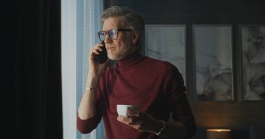 Kırmızı balıkçı yaka kazaklı ve gözlüklü şık Olgun Adam Parlak Otel Odası 'nda Panoramik Pencere kenarında durup telefonda konuşurken kahvesini yudumluyor. Kavram Sabah Rutini, İş Seyahati, Uzaktan İletişim.