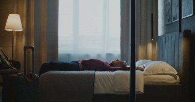 Olgun bir iş adamı, uzun bir iş seyahatinden sonra şık bir otel odasına girer. Yorgun ama rahatlamış, Erkek ceketini çıkarıp beyaz yatağa uzanıyor. Seyahat Yorgunluğu, Rahatlık ve Modern Yaşam Tarzı kavramı.