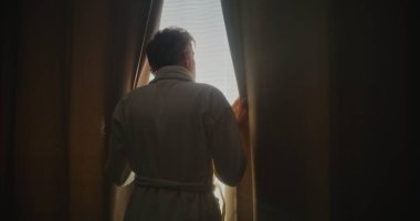 Gümüş Saçlı Adam Stylish Otel Odasında Yumuşak Güneş Işığından Hoşlanıyor. Adam pencereden dışarı bakar, ağır perdeleri geri çeker ve gerilir. Seyahat ederken rahatlama, duygusal refah kavramı. Arka Görünüm.