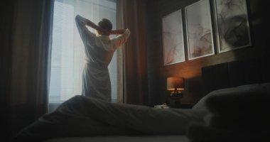 Modern Otel Odasında Yeni Gün 'e Hoş Geldiniz. Pijama ve bornoz giyen adam, güneşli pencereden dışarı bakarken yataktan çıkar ve gerilir. Rahatlama kavramı, seyahat ederken, duygusal refah....