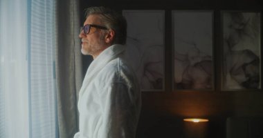 Yumuşak Beyaz Bornozlu Olgun Adam Cozy Otel Odasındaki Perdeleri Açar, Sıcak Gün Işığına İzin Verir. Huzurlu Gülümseme, Gün be gün sükuneti yansıtır. Düşünceli Seyahat, Sabah Dengesi ve İç Uyum kavramı.