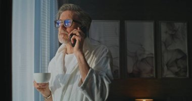 Otel Bornozunda Kahve İle İş Görüşmesi Yöneticisi. Beyaz Otel Bornozlu ve Gözlüklü Olgun Adam Sunny Window 'un yanında duruyor, Telefonla Nişanlanırken Kahve yudumluyor. Uzak Çalışma Yaşam Biçimi.