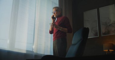 Kırmızı Balıkçı yaka kazaklı Olgun Adam ve Gözlüklü Stands by Large Window, Modern Otel Odasında Telefon Çağrısına Bağlıyken Kahve ya da Çay İçen Adam. Konsept İş İletişimi, Uzaktan Çalışma Hayatı. El bilgisayarı.