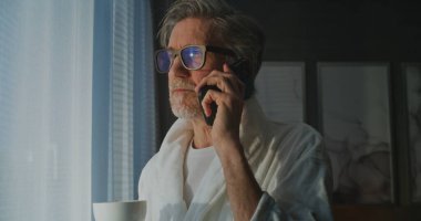 Beyaz Bornozlu ve Gözlüklü Olgun İşadamı Bright Otel Penceresinin yanında duruyor, Telefonda ve Kahve Kupasında Konuşuyor. Morning Light Modern Uzaktan Çalışma, Konfor ve İş Yaşam Tarzını Sembolize Ediyor.