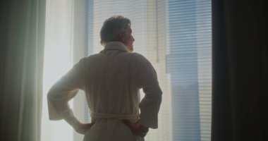 Olgun adam pencereden dışarı bakar, ağır perdeleri geri çeker ve gerilir. Parlak güneş ışığıyla dolu modern bir otel odası. Rahatlama, seyahat, günaydın, duygusal refah kavramı. Arka Görünüm.