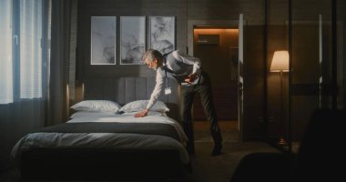 Resmi Takım Elbiseli Otel Yöneticisi Tablet Fotoğrafı kullanarak Lüks Otel Odasını Denetliyor ve Yatak Düzenlemesini Değerlendiriyor. Konukseverlik Standartları, Kalite Kontrolü, Profesyonel Yönetim.