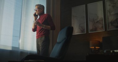 Gri saçlı ve gözlüklü kendine güvenen işadamı telefon görüşmesi yaparken Büyük Pencere 'nin yanında kahve içiyor. İş hayatı dengesi, uzaktan çalışma, iş seyahati, modern dijital yaşam tarzı. El bilgisayarı.