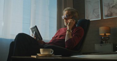 Kucağında tabletle Modern Otel Odası 'nda oturan Gri Saçlı İşadamı telefonda kahvaltı servisi olarak konuşuyor. Uzaktan Çalışma, İş Seyahati ve Dijital İletişim Konsepti. El bilgisayarı.