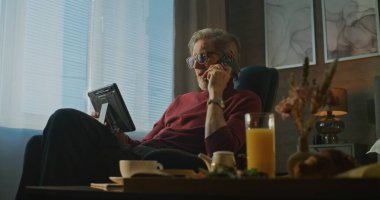 Kucağında tabletle Modern Otel Odası 'nda oturan Gri Saçlı İşadamı telefonda kahvaltı servisi olarak konuşuyor. Uzaktan Çalışma, İş Seyahati ve Dijital İletişim Konsepti. El bilgisayarı.