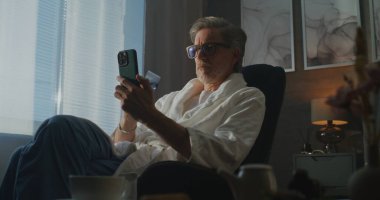 Otel Bornozundaki Olgun Adam Rahat Sabah Işığında Güvenli Çevrimiçi Ödeme İçin Akıllı Telefon ve Kredi Kartı Kullanıyor. Dijital Yaşam Tarzı ve Seyahat Ederken Uzaktan İşlemler Uygulaması. El bilgisayarı.