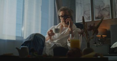 Otel odasındaki bornozda uzaktan kumandayla çalışan Yönetici Sabahı. Kol sandalyesinde oturan, telefonla konuşan, odada dinlenen gri saçlı adam. Bütünleşik İş ve Kişisel Yaşam kavramı.