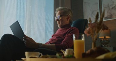 Gri Saçlı Adam Otel Sandalyesinde Rahatça Oturur, Kahvesini yudumlar ve Sabah Işığında Dizüstü Bilgisayarda Yazar ve Sıcak Atmosfer. Uzaktan Çalışma, İş Seyahati ve Dijital Esneklik kavramı. El bilgisayarı.