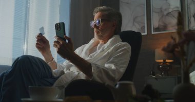 Otel Bornozundaki Olgun Adam Rahat Sabah Işığında Güvenli Çevrimiçi Ödeme İçin Akıllı Telefon ve Kredi Kartı Kullanıyor. Dijital Yaşam Tarzı ve Seyahat Ederken Uzaktan İşlemler Uygulaması. El bilgisayarı.